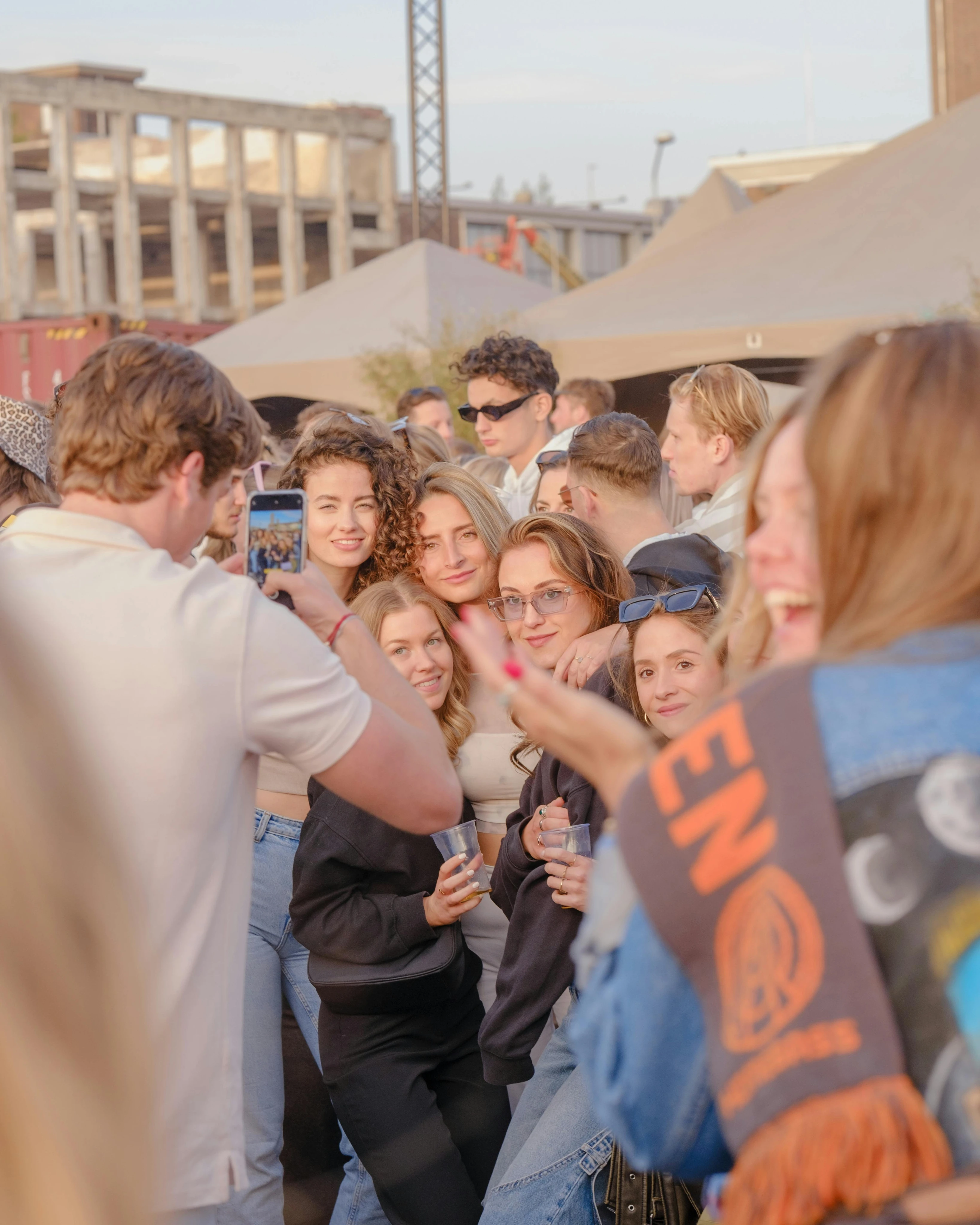Groep jonge volwassenen die lachen en poseren voor een foto op een buitenevenement, met een man die een telefoon vasthoudt om het moment vast te leggen. De setting bevat tenten en een gedeeltelijk gebouwd of verlaten gebouw op de achtergrond, wat zorgt voor een ontspannen en levendige sfeer.